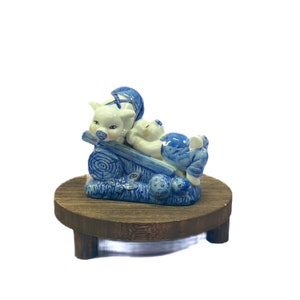 May include: A blue and white porcelain figurine of two pigs, one pushing a wheelbarrow, on a wooden base. The pigs are wearing blue and white clothing.