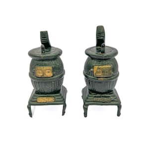 May include: Two miniature black cast iron wood stoves with gold accents. The stoves have a round body with a door on the front and a chimney on top. The stoves are sitting on a white surface.