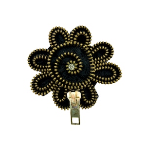 Op de afbeelding: Een zwarte, bloemvormige broche gemaakt van goudkleurige ritsen met een heldere strasssteen in het midden. De broche heeft een goudkleurige rits-trekker.
