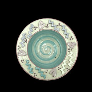 May include: A handmade ceramic bowl with a light green exterior and a turquoise interior. The rim features zigzag and heart-shaped designs in gray and pink. The bowl's interior has a spiral pattern.