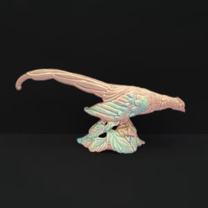May include: A ceramic pheasant figurine in shades of pink, blue, and green. The bird is perched on a base of stylized leaves. The pheasant has detailed feather patterns and a curved tail, set against a black background.
