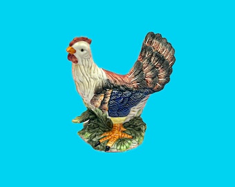 May include: A ceramic rooster figurine with a white, red, and brown body, blue and black wings, and yellow feet. The rooster is perched on a green base with leaf details. The background is a solid light blue.