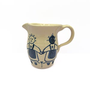 May include: A cream-colored ceramic pitcher with a blue and black hand-painted design of two figures holding hands. The figures are wearing dresses and have simple facial features.