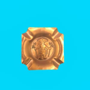 May include: A square, bronze-colored ashtray with a central embossed design of a figure holding tools. The ashtray has four corner cutouts for resting cigarettes. The background is a solid, bright blue.