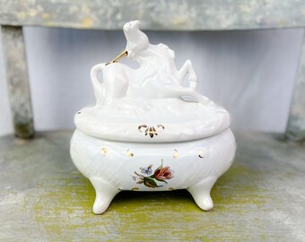 May include: White porcelain trinket box with a unicorn lid and floral accents. The box has three legs and a gold trim.