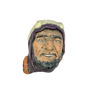 May include: A ceramic bust of a Middle Eastern man with a weathered appearance. The sculpture features a tan face, a dark beard, and a purple headdress with a cream-colored top. The man's expression is a slight smile.