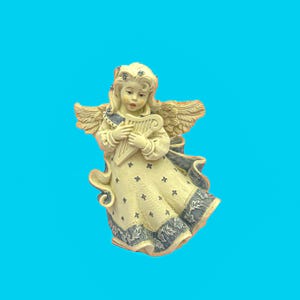 May include: A cream-colored angel figurine with blonde hair and feathered wings. The angel is holding a harp and wearing a dress with blue floral accents. The figurine has a detailed design and is set against a bright blue background.