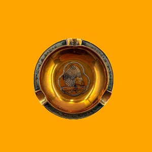 May include: A vintage ashtray with a gold-colored interior and a dark rim. The center features a detailed relief of a Native American chief. The ashtray has three notches for resting cigarettes.
