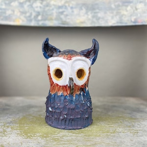 May include: A ceramic owl figurine with blue, purple, red, and white paint. The owl has large, round eyes with yellow pupils.