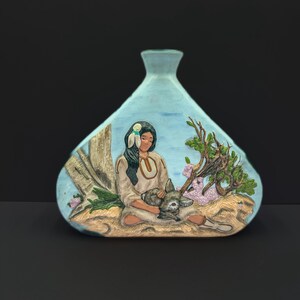 May include: A light blue, teardrop-shaped vase with a relief scene of a Native American woman holding a small animal. The woman is wearing a white dress and has long black hair. The scene includes a tree and flowers.
