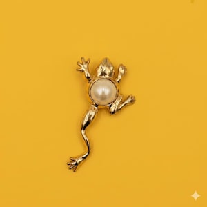 May include: A gold-toned metal brooch shaped like a frog with a smooth, round, white faux pearl set in the center of its body. The frog is depicted in a leaping pose with extended limbs and detailed feet, featuring a polished, reflective surface.