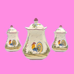 May include: Three ceramic canisters with lids. The canisters are off-white with a rooster design in yellow, blue, and red. The lids have a floral design and a small, round knob. The canisters are decorated with green and brown foliage.