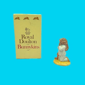 May include: A Royal Doulton Bunnykins figurine of a kneeling bunny in blue and white striped pajamas, with a brown bunny toy. The figurine is on a yellow base. The box next to it reads "Royal Doulton Bunnykins".