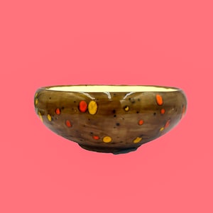 May include: A brown ceramic bowl with a speckled design of orange and yellow dots. The interior of the bowl is a light yellow color. The bowl is set against a pink background.