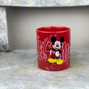 Pu&ograve; includere: Tazza in ceramica rossa con un design di Topolino bianco. La tazza ha le parole "Mickey Mouse" scritte in bianco, in un font stilizzato.