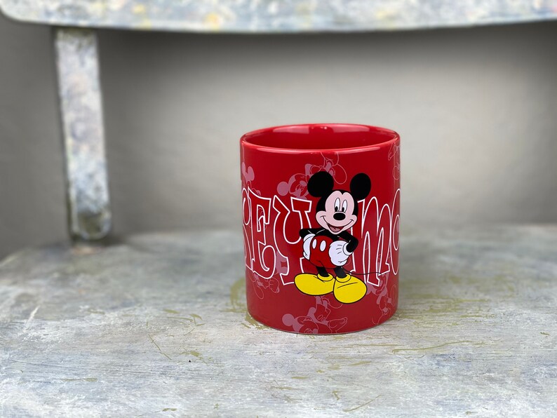 Pu&ograve; includere: Tazza in ceramica rossa con un design di Topolino bianco. La tazza ha le parole "Mickey Mouse" scritte in bianco, in un font stilizzato.