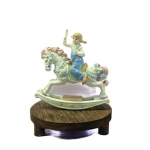 May include: A ceramic rocking horse figurine with a child rider, painted in pastel colors. The horse has a light green body with pink and blue accents. The child wears blue overalls and is waving. The figurine sits on a small, round wooden stand.