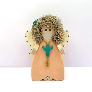 May include: A wooden angel figurine with a peach-colored dress and cream-colored wings. The angel has a teal cactus pendant and moss hair. The wings are decorated with teal polka dots. A teal bow adorns the top of the head.