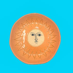 May include: A handmade ceramic plate with a sun design. The plate is orange with a cream-colored face in the center, featuring black eyes and a simple nose and mouth. The sun's rays are etched around the face.