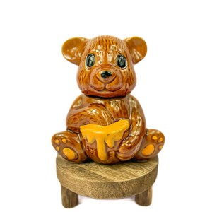 May include: A brown ceramic bear figurine sitting on a wooden stool. The bear is holding a honey pot with dripping honey.