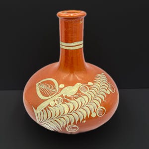 May include: A terracotta vase with a rounded body and a narrow neck. The vase is decorated with a white bird and fern design.