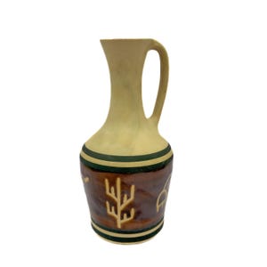 May include: A small, beige ceramic pitcher with a brown and green band around the middle. The pitcher has a design of a tree and other geometric shapes in brown on the front.
