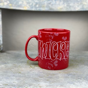 Pu&ograve; includere: Tazza in ceramica rossa con un design di Topolino bianco. La tazza ha un contorno bianco della testa di Topolino e la parola "Mickey" in un font stilizzato.
