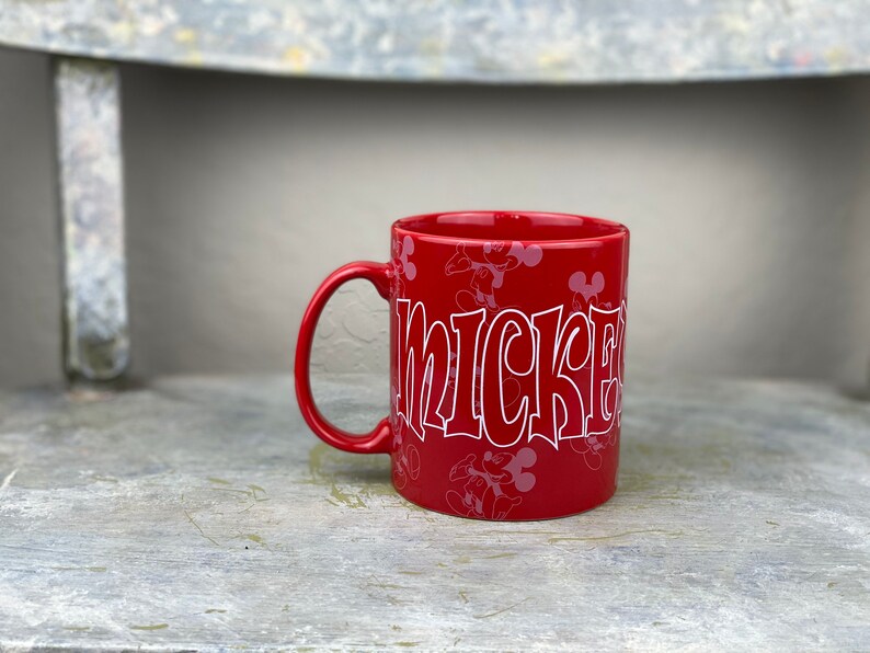 Pu&ograve; includere: Tazza in ceramica rossa con un design di Topolino bianco. La tazza ha un contorno bianco della testa di Topolino e la parola "Mickey" in un font stilizzato.