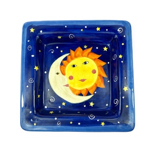 May include: A blue ceramic square dish with a white rim and a painted design of a yellow sun and a white crescent moon on a blue background with white stars.