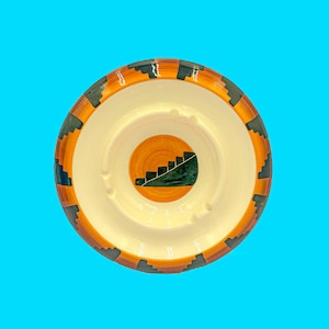 May include: A ceramic ashtray with a cream interior and an orange and black patterned border. The centre has an orange circle with a green and white staircase design. The ashtray has four cigarette rests.