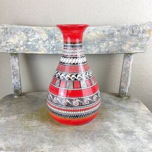 May include: A red ceramic vase with a geometric pattern in black and white. The vase has a wide base and a narrow neck.