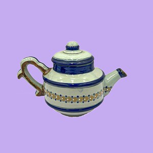 May include: A ceramic teapot with a lid, handle, and spout. The teapot is white with blue and brown decorative bands and accents. The handle is brown and the spout is blue. The teapot is on a purple background.
