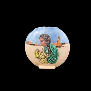 May include: A round, cream-colored ceramic piece featuring a Native American woman in a green dress holding a baby wrapped in yellow. The background depicts a desert landscape with a blue sky and a teepee.