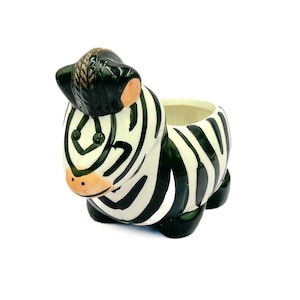 May include: A ceramic planter shaped like a zebra with black and white stripes. The planter has a small opening at the top for planting.