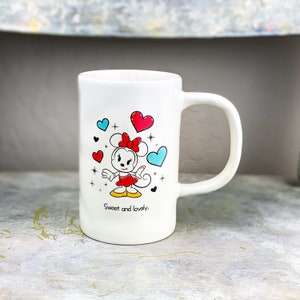 Pu&ograve; includere: Tazza in ceramica bianca con un'immagine di Minnie Mouse in un vestito rosso e un fiocco. La tazza ha cuori blu e rossi e stelle intorno all'immagine. Il testo "Sweet and lovely." &egrave; stampato sul fondo della tazza.