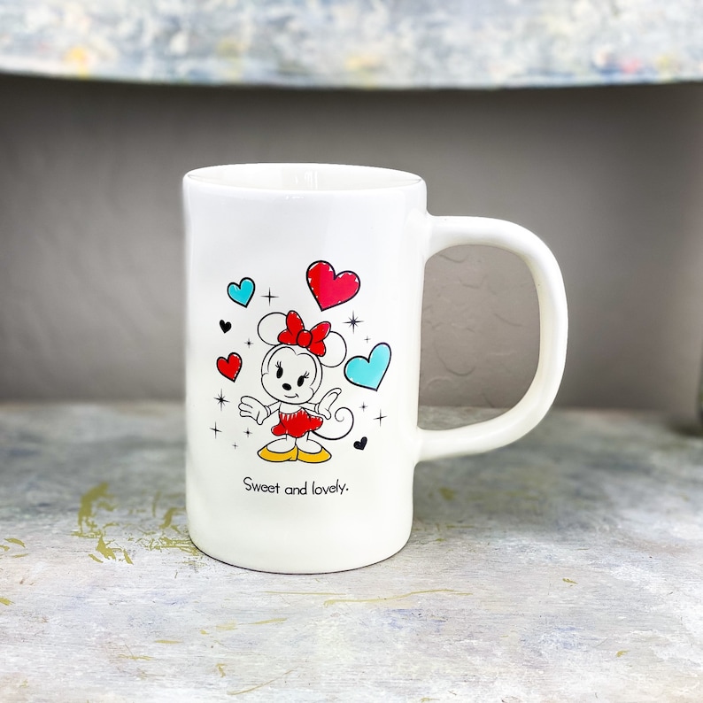 Pu&ograve; includere: Tazza in ceramica bianca con un'immagine di Minnie Mouse in un vestito rosso e un fiocco. La tazza ha cuori blu e rossi e stelle intorno all'immagine. Il testo "Sweet and lovely." &egrave; stampato sul fondo della tazza.