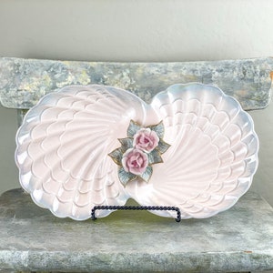 May include: A pink ceramic serving dish shaped like two seashells with a raised floral design in the center. The dish is on a black metal stand.