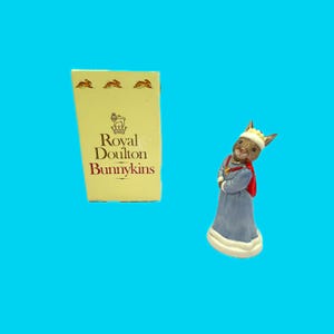 May include: A Royal Doulton Bunnykins figurine of a rabbit wearing a blue robe with a red cape and a gold crown. The figurine is next to a yellow box with the words "Royal Doulton Bunnykins" printed on it.