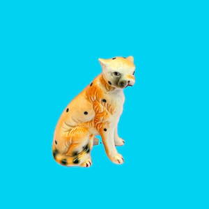 May include: A ceramic figurine of a seated leopard with a cream-colored face and body, orange spots and markings, and black spots. The leopard has a detailed face and is set against a bright blue background.