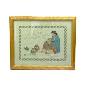 May include: A framed cross-stitch embroidery depicting a woman in traditional clothing sitting with pottery in a desert landscape. The embroidery is signed "mja 2004".