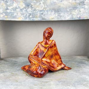 May include: A ceramic figurine of a seated person with a draped robe. The sculpture has a glossy finish with shades of brown and yellow. The figure is in a meditative pose, with the head slightly bowed.