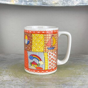 May include: A white ceramic mug with a patchwork design featuring a hot air balloon, sailboat, rainbow, and house. The mug has an orange rim and handle.