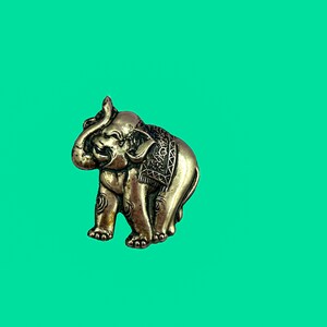 May include: A detailed, antique-style elephant figurine in a metallic silver color. The elephant has intricate carvings on its body and is set against a bright teal background. The trunk is raised, and the elephant appears to be in a standing position.