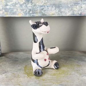 May include: A ceramic cow figurine with black and white spots, wearing a black and white hat and a pink tongue sticking out. The cow has red buttons on its belly and is sitting on a wooden surface.