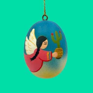 May include: A hand-painted, egg-shaped ornament with a blue background. It features a figure with angel wings and a red dress, holding a cactus in a pot. The ornament has a gold string for hanging.