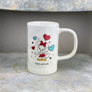 Pu&ograve; includere: Tazza in ceramica bianca con un'immagine di cartone animato di Minnie Mouse con un vestito rosso e un fiocco. La tazza ha cuori blu e rossi e il testo "Sweet and lovely."