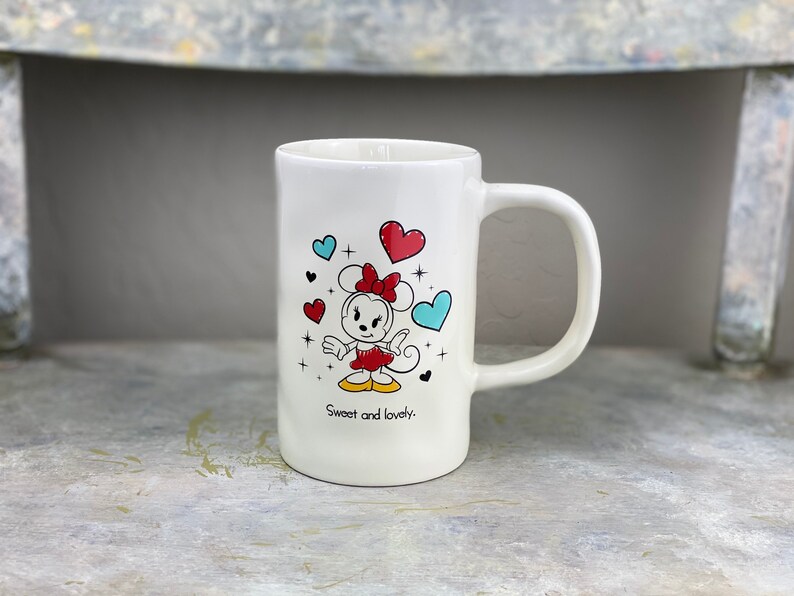 Pu&ograve; includere: Tazza in ceramica bianca con un'immagine di cartone animato di Minnie Mouse con un vestito rosso e un fiocco. La tazza ha cuori blu e rossi e il testo "Sweet and lovely."