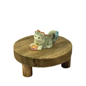 May include: A gray and white cat figurine sitting on a wooden table with a pink bowl that says "Kitty" in front of it. The cat is wearing a blue and white bow on its tail.