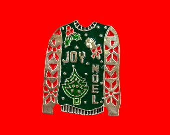 Può includere: Spilla a forma di maglione natalizio con base verde e finiture argentate. La spilla presenta le parole "JOY" e "NOEL", un albero di Natale, agrifoglio e un omino di pan di zenzero. Lo sfondo è rosso.