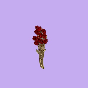 May include: A gold-toned brooch featuring a bouquet of red roses. The roses are clustered together, with detailed petals and a realistic appearance. The stems and leaves are also gold, creating a cohesive and elegant design. The brooch is set against a lavender background.